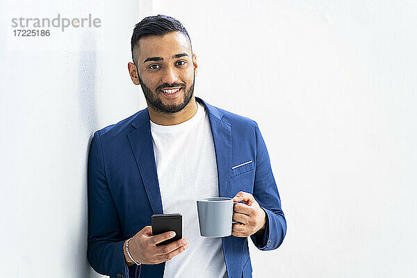 Männlicher Unternehmer mit Kaffeetasse und Smartphone in der Hand  während er sich im Büro an die Wand lehnt
