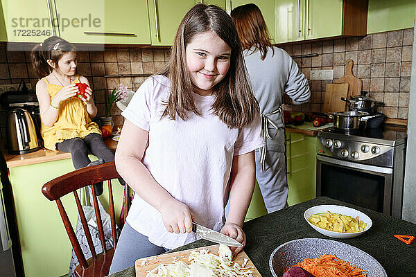 Lächelndes Mädchen  das Kohl schneidet  während Mutter und Schwester im Hintergrund Essen zubereiten