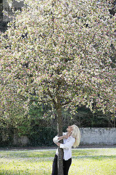 Fröhliche Frau mit umarmendem Baum im Park während des Frühlings