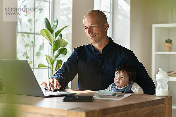 Männlicher Unternehmer arbeitet am Laptop  während er sein Baby im Home Office hält