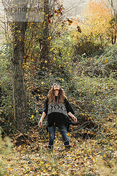 Frau betrachtet Herbstblätter in der Luft  während sie in einem Wald im Urlaub steht