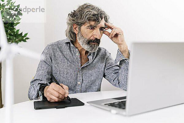 Konzentrierter älterer Geschäftsmann mit Grafiktablett am Laptop  während er im Büro sitzt