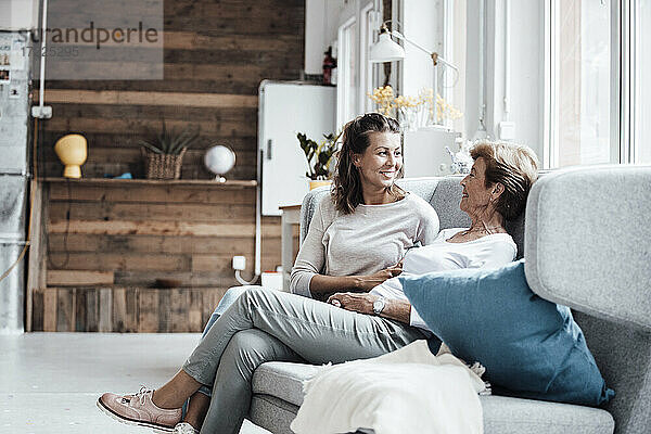 Lächelnde junge Frau im Gespräch mit ihrer Großmutter  während sie sich zu Hause auf dem Sofa entspannt