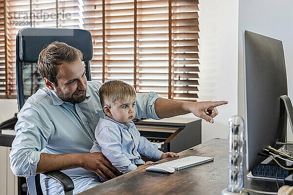 Lächelnder Vater  der auf einen Computer zeigt  während sein Sohn zu Hause auf dem Schoß sitzt