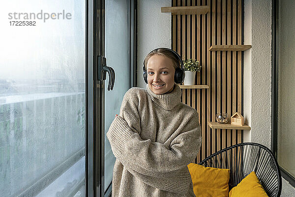 Junge lächelnde Frau mit Kopfhörern  die mit verschränkten Armen vor einem Fenster zu Hause steht