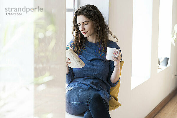 Schöne Frau liest ein Buch  während sie einen Kaffee am Fenster im Wohnzimmer trinkt