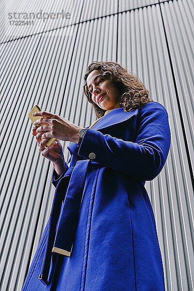 Geschäftsfrau im Trenchcoat  die ein Mobiltelefon benutzt  während sie vor einer grauen Wand steht