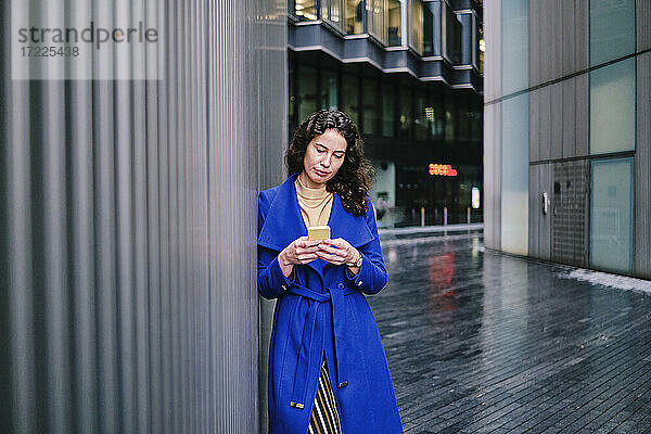 Geschäftsfrau in blauem Trenchcoat arbeitet an einem Mobiltelefon und lehnt sich an die Wand