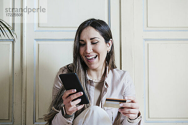 Glückliche Frau  die ein Smartphone benutzt und eine Kreditkarte zu Hause hält