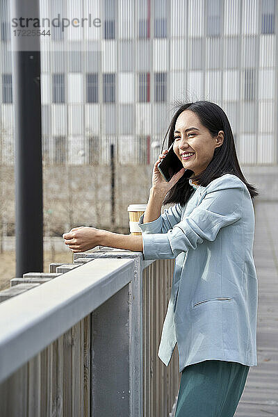 Lächelnde Geschäftsfrau  die mit ihrem Handy telefoniert und sich auf ein Geländer stützt