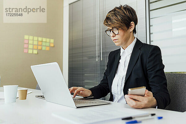 Geschäftsfrau  die einen Laptop benutzt und ein Mobiltelefon am Schreibtisch im Büro hält