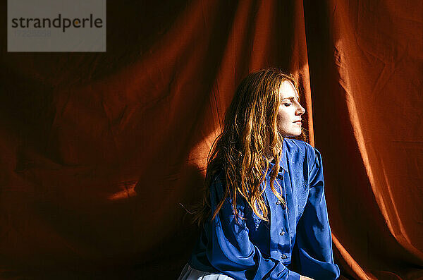 Junge Frau mit geschlossenen Augen vor einem roten Tuch im Sonnenlicht sitzend