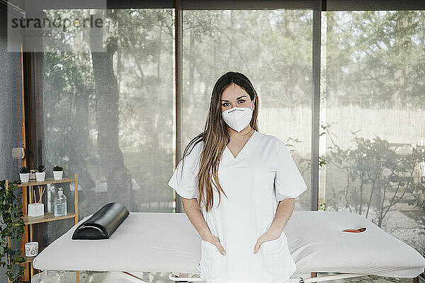 Ärztin mit Händen in den Taschen vor einem Massagetisch in einer Klinik während einer Pandemie