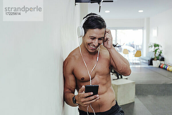 Männlicher Sportler stellt Kopfhörer ein  während er im Fitnessstudio ein Mobiltelefon benutzt