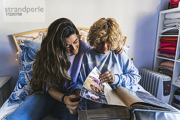 Lächelnde Tochter schaut sich ein Fotoalbum an  während sie mit ihrer Mutter im Schlafzimmer sitzt