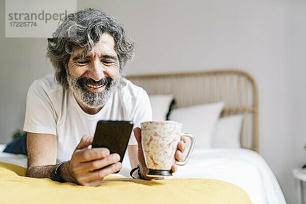 Glücklicher älterer Mann  der ein Smartphone benutzt und einen Kaffeebecher hält  während er zu Hause auf dem Bett liegt