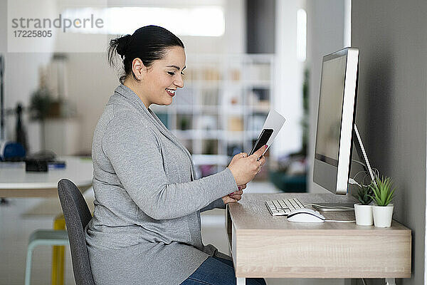 Lächelnde Unternehmerin  die ein Mobiltelefon benutzt und ein Dokument im Büro zu Hause hält