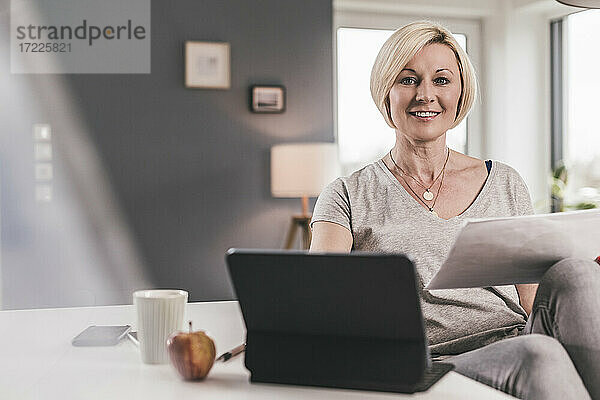 Lächelnde Frau  die Papier hält  während sie mit einem digitalen Tablet auf einem Tisch zu Hause sitzt