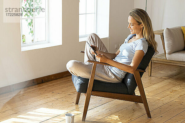 Lächelnde Frau  die ein digitales Tablet benutzt  während sie auf einem Stuhl im Wohnzimmer sitzt