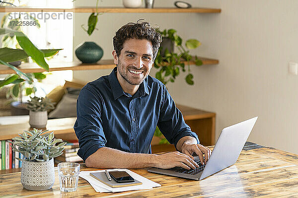 Glücklicher männlicher Freiberufler  der von zu Hause aus über seinen Laptop arbeitet