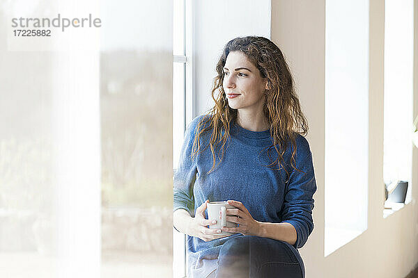 Junge Frau hält Kaffeetasse  während sie am Fenster im Wohnzimmer sitzt