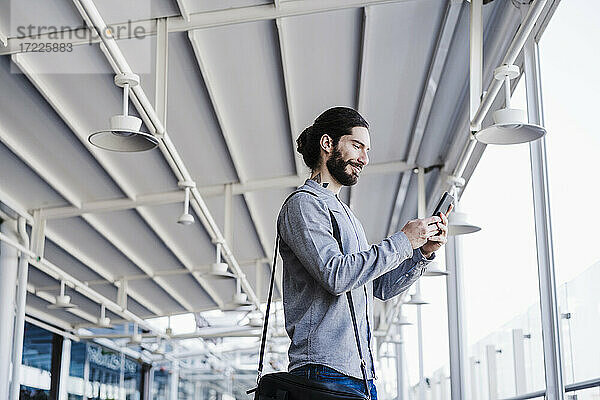 Lächelnder Geschäftsmann mit Laptoptasche  der ein Mobiltelefon auf dem Balkon eines Büros benutzt