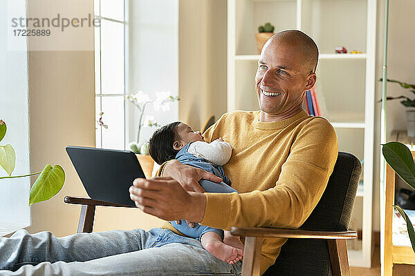 Glücklicher Vater hält schlafendes Baby und digitales Tablet  während er zu Hause wegschaut