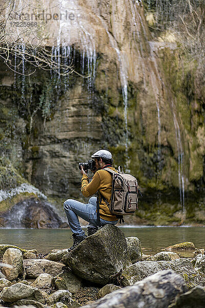 Männlicher Wanderer  der auf einem Felsen am See im Wald sitzt und fotografiert