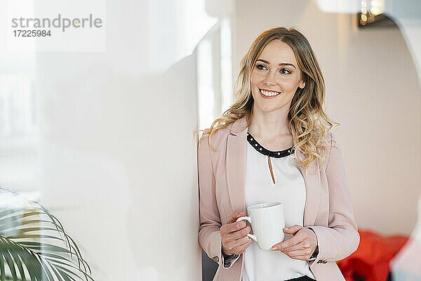 Lächelnde weibliche Fachkraft  die wegschaut und einen Kaffeebecher im Büro hält