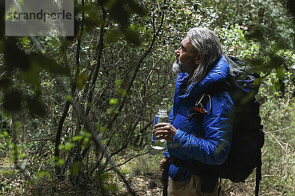 Älterer männlicher Forscher betrachtet Pflanzen und hält eine Wasserflasche im Wald