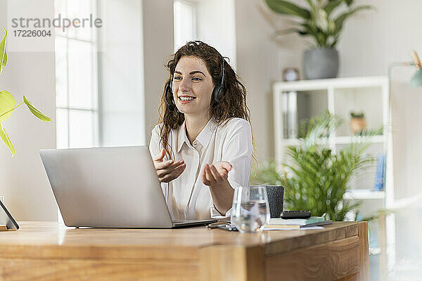 Lächelnde Frau gestikuliert  während sie im Büro zu Hause über Kopfhörer spricht