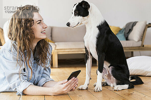Lächelnde Frau mit Mobiltelefon  die einen Jack Russell Terrier betrachtet  während sie im Wohnzimmer auf dem Boden liegt