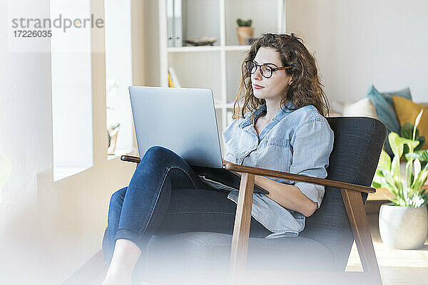 Frau  die einen Laptop benutzt und auf einem Sessel im Wohnzimmer sitzt