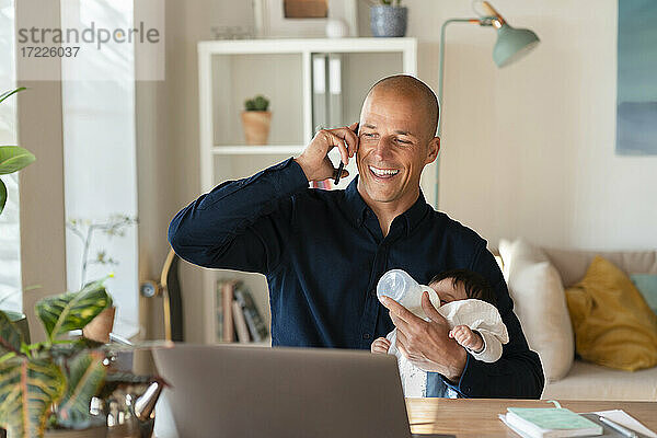 Lächelnder Vater  der mit seinem Smartphone telefoniert  während er seiner Tochter zu Hause Milch gibt
