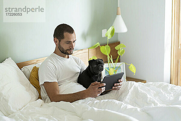 Junger Mann  der ein digitales Tablet mit einem Mops-Hund benutzt  während er zu Hause auf dem Bett sitzt