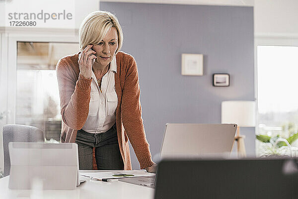 Weibliche Fachkraft  die auf einen Laptop schaut  während sie über ein Mobiltelefon im Heimbüro diskutiert