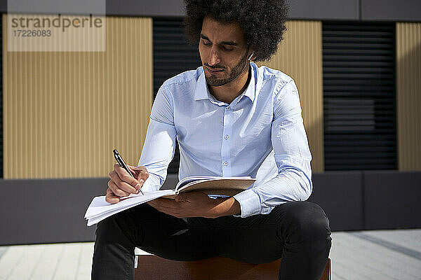 Afro-Mann schreibt in ein Buch  während er auf einem Stuhl auf der Straße sitzt