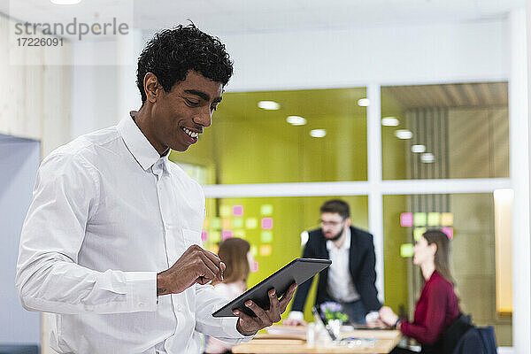 Lächelnder Geschäftsmann  der ein digitales Tablet benutzt  mit Kollegen im Hintergrund im Büro