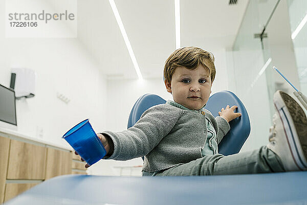 Kleinkind  das mit einer Tasse spielt  während es auf einem Stuhl in der Zahnarztpraxis sitzt