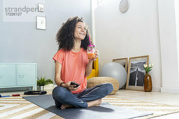 Fröhliche Frau beim Safttrinken mit Handy auf der Matte im Wohnzimmer