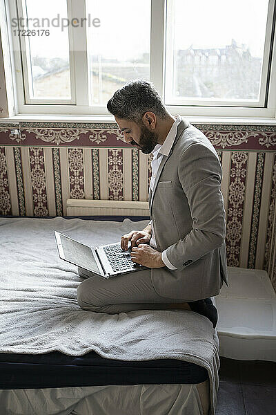 Männlicher Unternehmer  der einen Laptop benutzt  während er zu Hause auf dem Bett sitzt