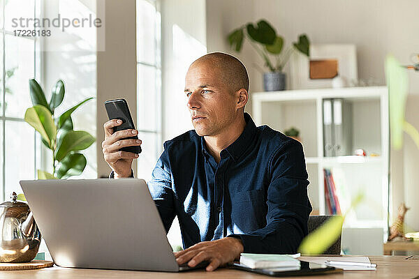 Männlicher Unternehmer arbeitet am Smartphone  während er zu Hause am Laptop sitzt