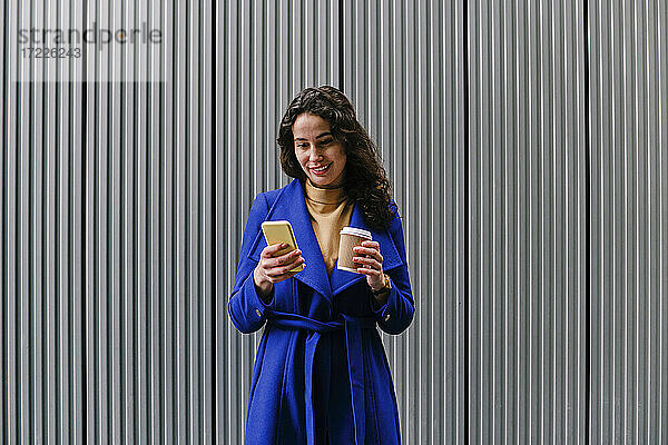 Lächelnde Geschäftsfrau  die einen Einweg-Kaffeebecher hält  während sie ein Mobiltelefon vor einer Wand benutzt