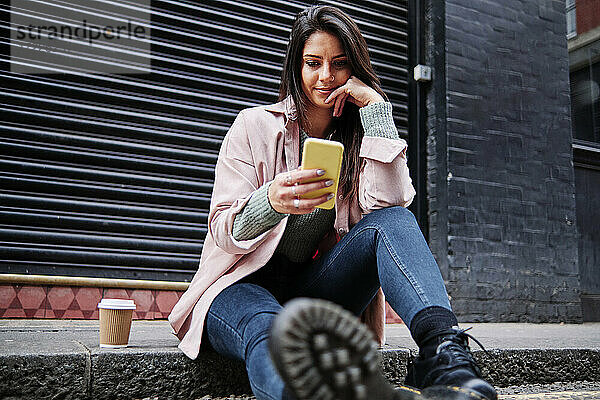 Schöne Frau mit Kaffeetasse und Mobiltelefon auf dem Bürgersteig sitzend