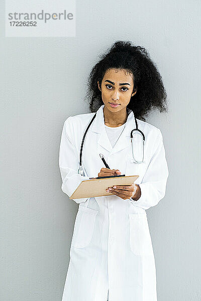 Selbstbewusste Ärztin  die ein Klemmbrett hält  während sie vor einer Wand steht