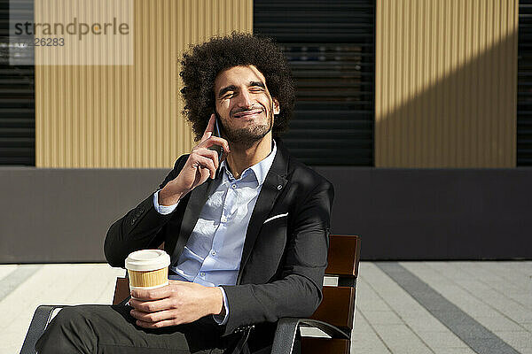 Lächelnder Afro-Mann  der mit seinem Smartphone spricht und eine Kaffeetasse hält  während er an einem sonnigen Tag auf einem Stuhl sitzt