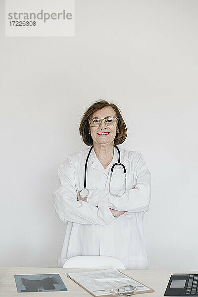 Lächelnde Ärztin mit verschränkten Armen vor einem Schreibtisch im Krankenhaus stehend