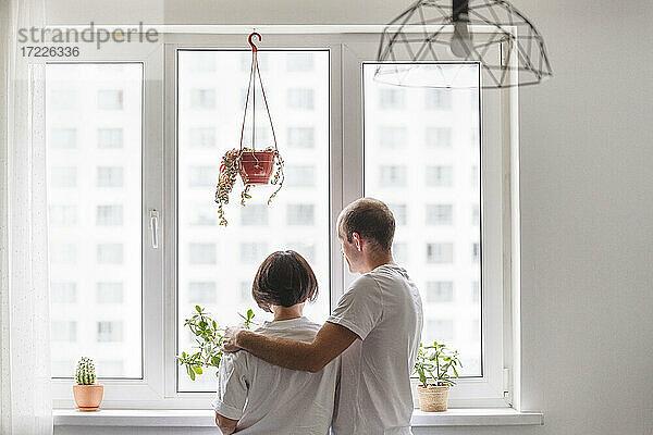 Ehemann und Ehefrau umarmen sich  während sie durch das Fenster zu Hause schauen