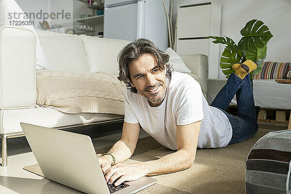 Nachdenklicher Mann mit Laptop  der wegschaut  während er zu Hause auf dem Boden liegt
