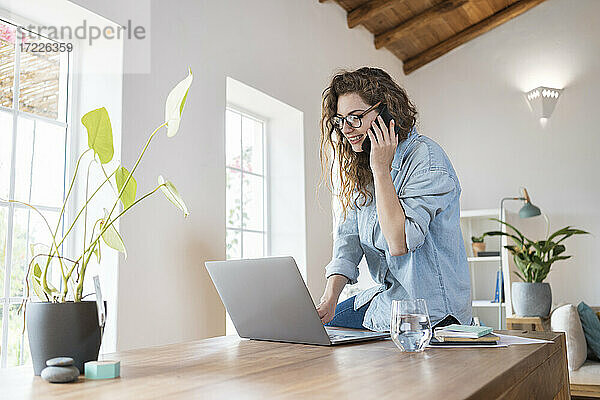 Lächelnde weibliche Fachkraft  die mit einem Smartphone telefoniert  während sie an einem Laptop am Schreibtisch arbeitet